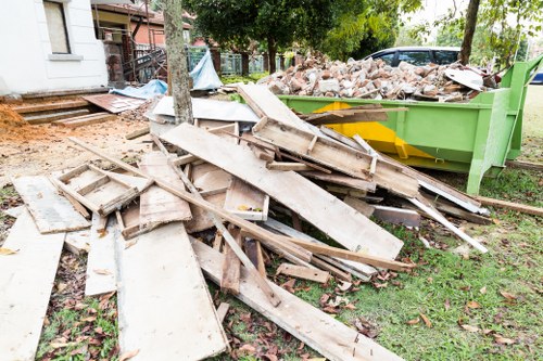 A pile of discarded wooden planks, some weathered and broken, is spread across a grassy area next to a green waste skip filled with a mixture of bricks and rubble. The wooden debris includes various sizes of planks, some with nails or rough edges, and are stacked haphazardly on the lawn, indicating recent clearing or renovation work. In the background, there are trees, a paved pathway, and part of a residential building, suggesting this scene is in a suburban garden or outdoor space. The waste removal process appears to involve the collection of construction debris, consistent with garden clearance or building renovation activities in Mitcham, as managed by Garden Clearance Mitcham, operating within the local area. Natural daylight illuminates the scene, emphasizing the textures of the wood and brick materials along with the surrounding greenery.