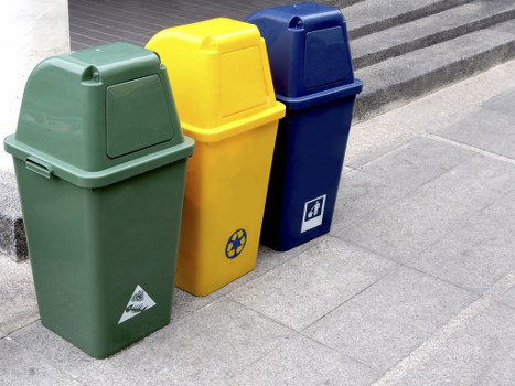 Three wheeled rubbish bins are positioned on a paved area beside a building entrance, likely on a residential or commercial street. The first bin on the left is green with a smooth, matte finish and a slightly domed lid. The middle bin is yellow, featuring a similar matte texture and a recycling symbol on its side, indicating its use for recyclable waste. The rightmost bin is dark blue, also with a matte surface and a small label or symbol on the front, possibly related to waste separation or recycling. All three bins are aligned closely together, standing upright on the grey pavement, just before a step or platform area. The background shows a portion of a dark surface, such as a concrete or stone step, with the bins positioned on a flat paved surface. The scene suggests a location where a waste collection service, like that offered by Garden Clearance Mitcham, might service local properties, fitting within the context of rubbish removal in Mitcham or nearby areas.