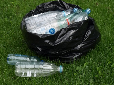 A black plastic rubbish bag positioned on a grassy surface, partially open and filled with several clear plastic bottles featuring blue and red caps. The bottles, varying in size, are visibly transparent, with some lying on the grass outside the bag, and others nestled securely inside. The grassy area appears well-maintained, with a mix of green blades, and the scene is illuminated by natural daylight, indicating an outdoor setting. The image showcases waste voluntary collected for disposal by a rubbish removal service, with the focus on the plastic bottles and the black bag used for rubbish containment, reflecting typical environmental waste disposal practices without specific location details. The scene visually emphasizes waste collection involving plastic bottles, aligning with rubbish removal activities in Mitcham or nearby areas, as represented by the context of the websiteGardenclearancemitcham.co.uk and the service page for garden clearance services.