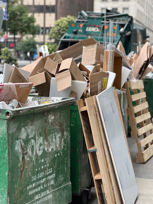 A large green industrial waste container filled with a variety of discarded cardboard boxes, paper packaging, and miscellaneous packaging materials. The cardboard boxes are in different sizes, some fl