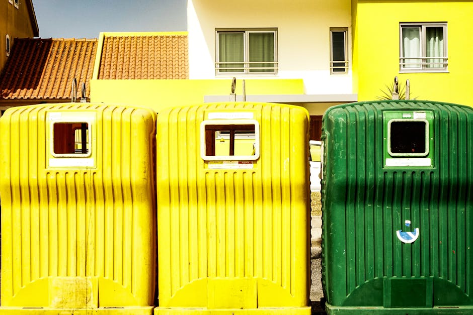 A large green industrial waste container filled with a variety of discarded cardboard boxes, paper packaging, and miscellaneous packaging materials. The cardboard boxes are in different sizes, some fl