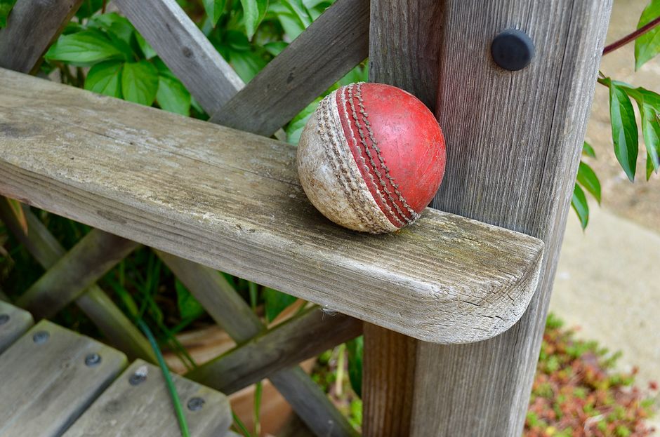 A weathered wooden garden bench with a rough, grayish finish, positioned outdoors amidst lush green foliage. Resting on the seat is a well-used, partially deflated red and white cricket ball with scra
